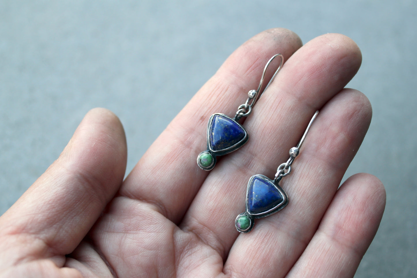 Silver earrings with blue and green stones held in a hand against a gray background