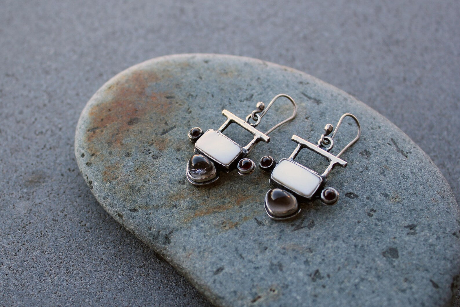 Handmade Armenian artisan sterling silver silversmith drop earrings featuring rectangular mother of pearl centers, accented by small round red garnets on the sides and teardrop-shaped grey obsidian at the base