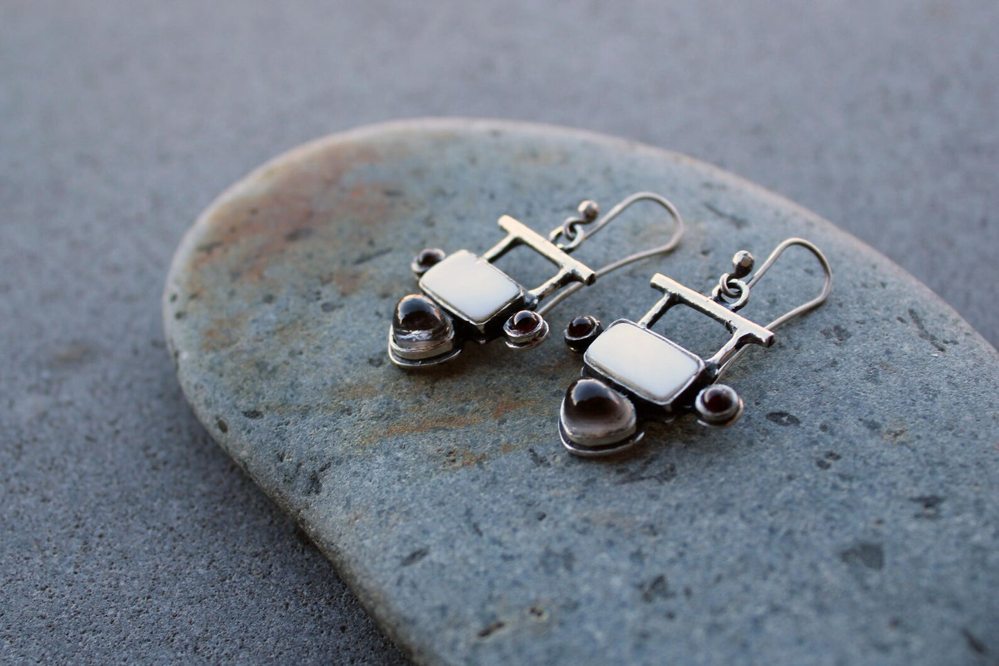 Handmade Armenian artisan sterling silver silversmith drop earrings featuring rectangular mother of pearl centers, accented by small round red garnets on the sides and teardrop-shaped grey obsidian at the base
