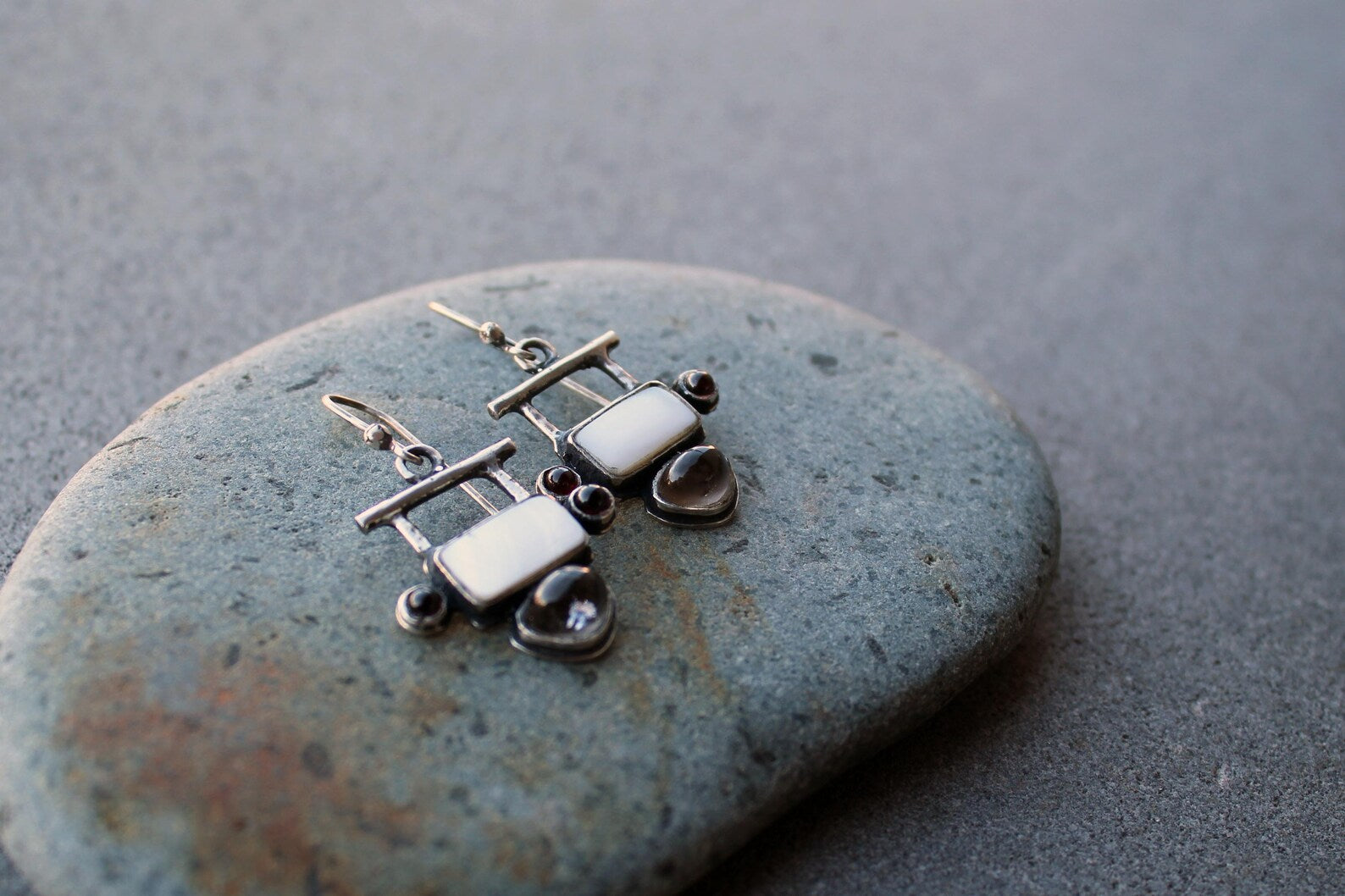 Handmade Armenian artisan sterling silver silversmith drop earrings featuring rectangular mother of pearl centers, accented by small round red garnets on the sides and teardrop-shaped grey obsidian at the base