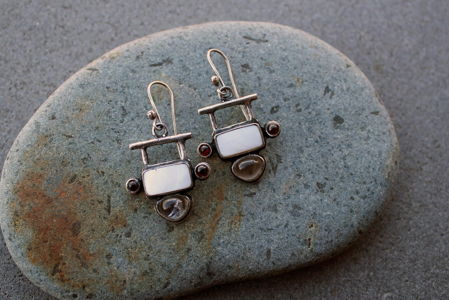 Handmade Armenian artisan sterling silver silversmith drop earrings featuring rectangular mother of pearl centers, accented by small round red garnets on the sides and teardrop-shaped grey obsidian at the base