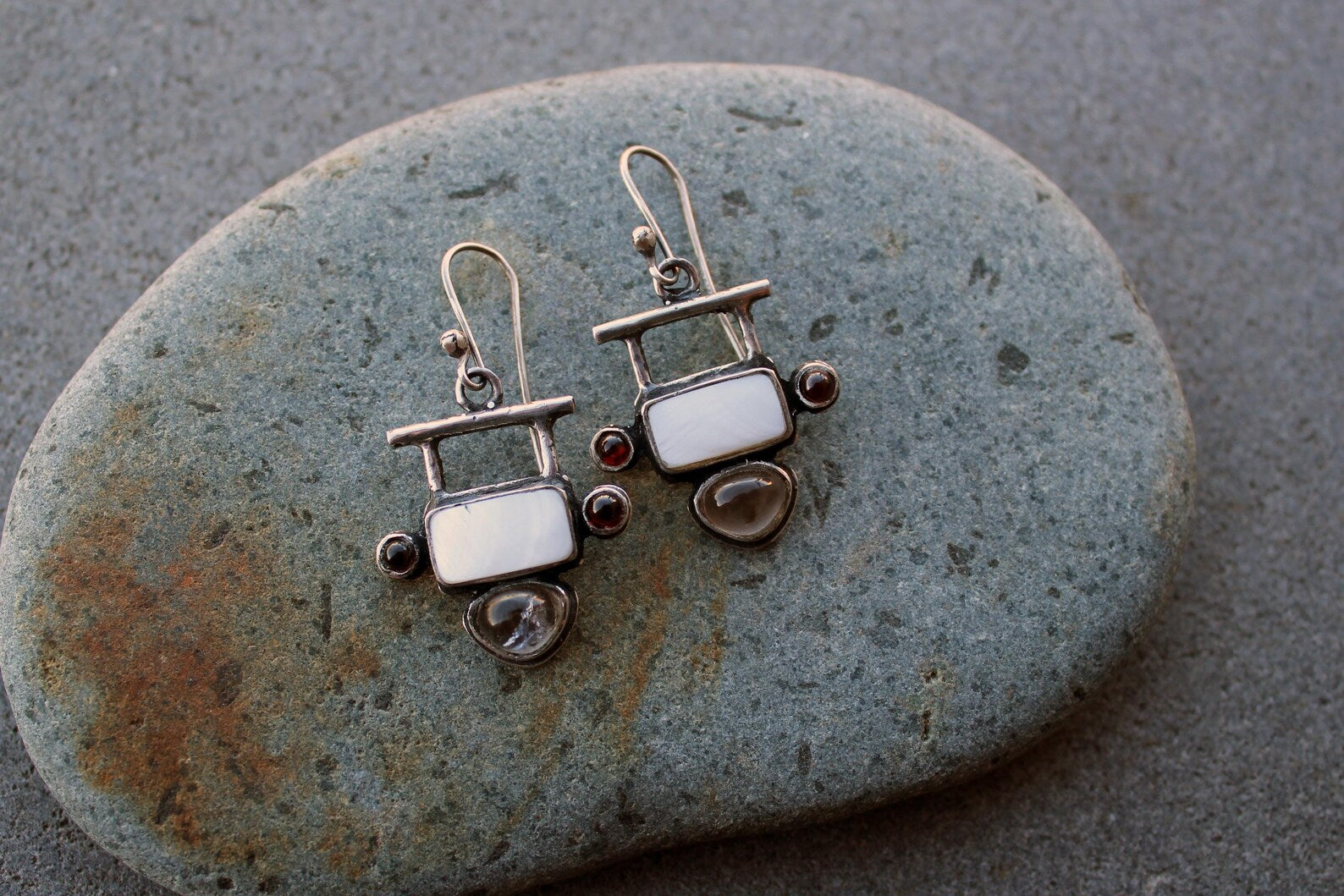 Handmade Armenian artisan sterling silver silversmith drop earrings featuring rectangular mother of pearl centers, accented by small round red garnets on the sides and teardrop-shaped grey obsidian at the base