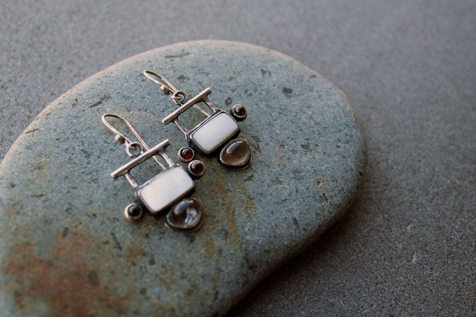 Handmade Armenian artisan sterling silver silversmith drop earrings featuring rectangular mother of pearl centers, accented by small round red garnets on the sides and teardrop-shaped grey obsidian at the base